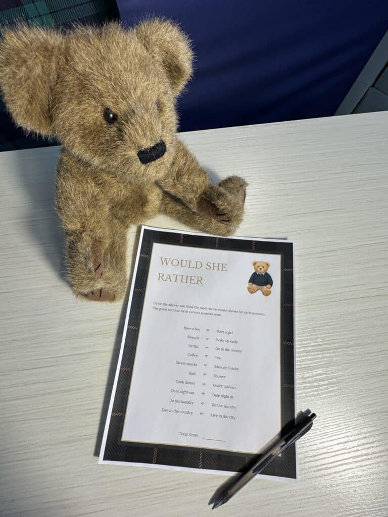 Game table with a teddy bear, pen, and a “Would She Rather?” baby shower game card featuring cute neutral graphics and multiple either‑or questions for guests to circle.